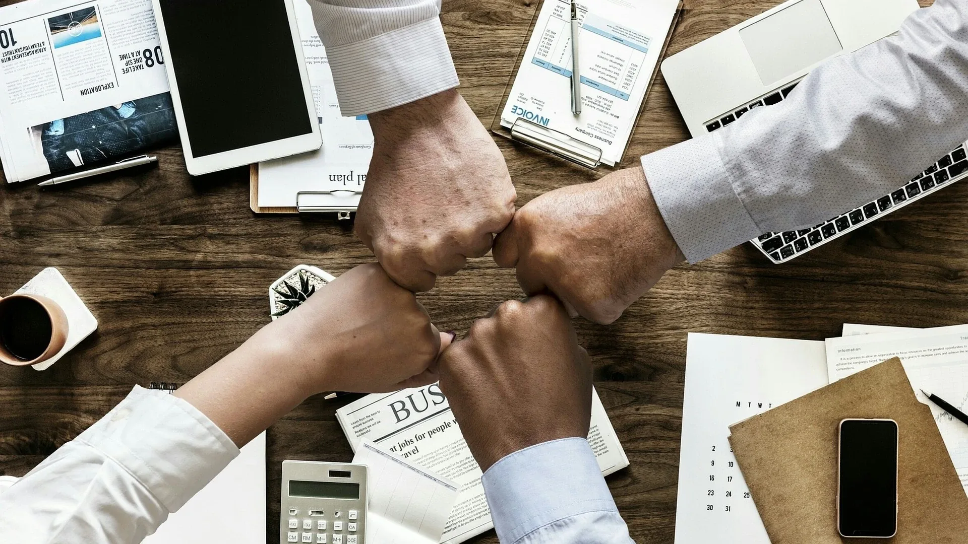 Four people bumping fists together over a shared desk of laptops, papers and notebooks